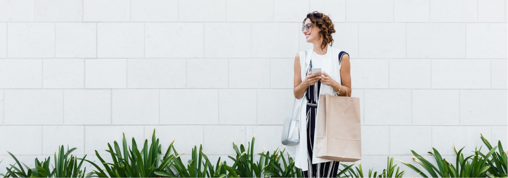 Woman standing in front of white bricks, smiling, and holding coffee and shopping bag